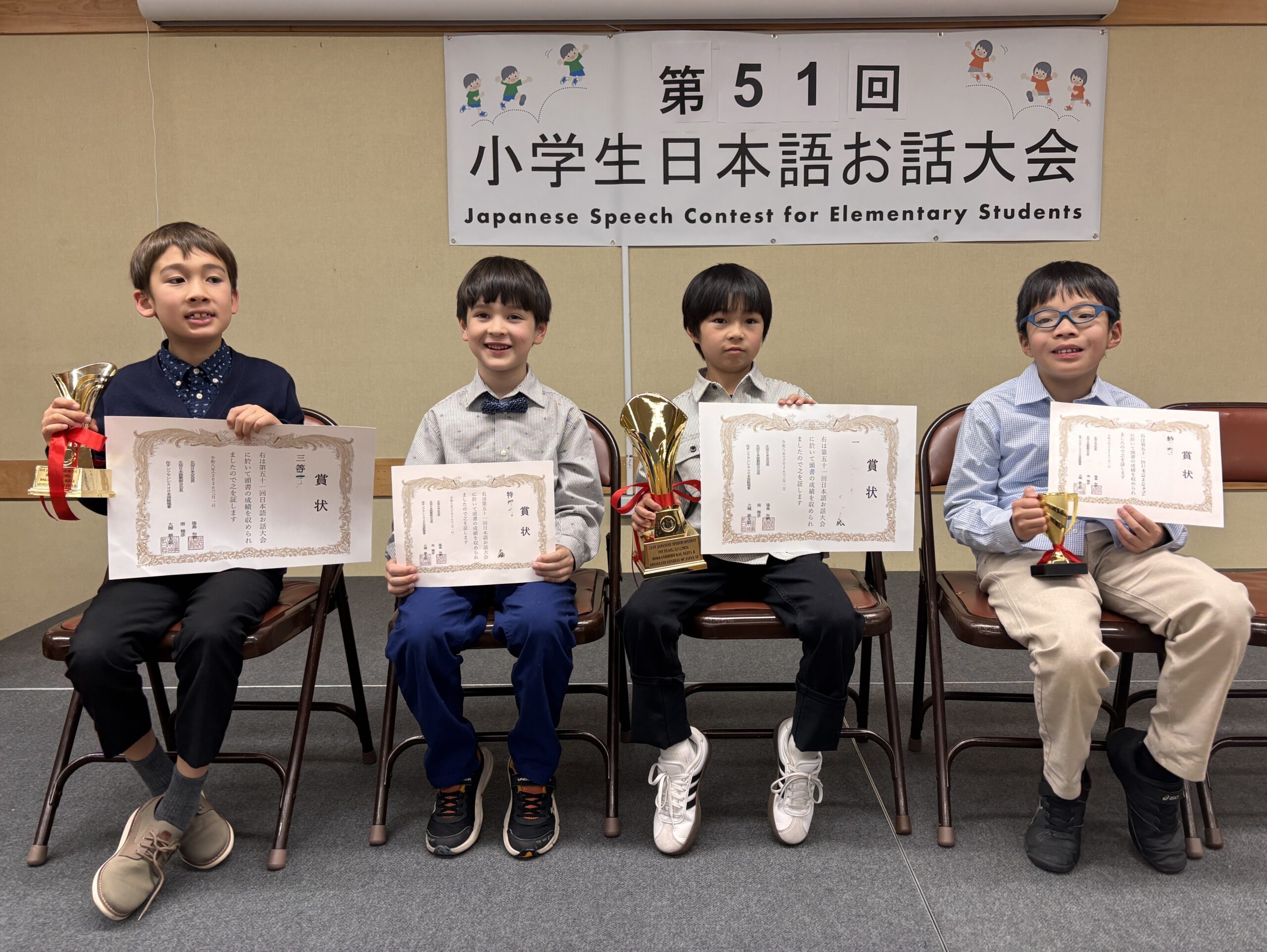 Four elementary school boys holding trophies and award certificates, sitting in front of a banner for the 51st Japanese Speech Contest for Elementary Students.